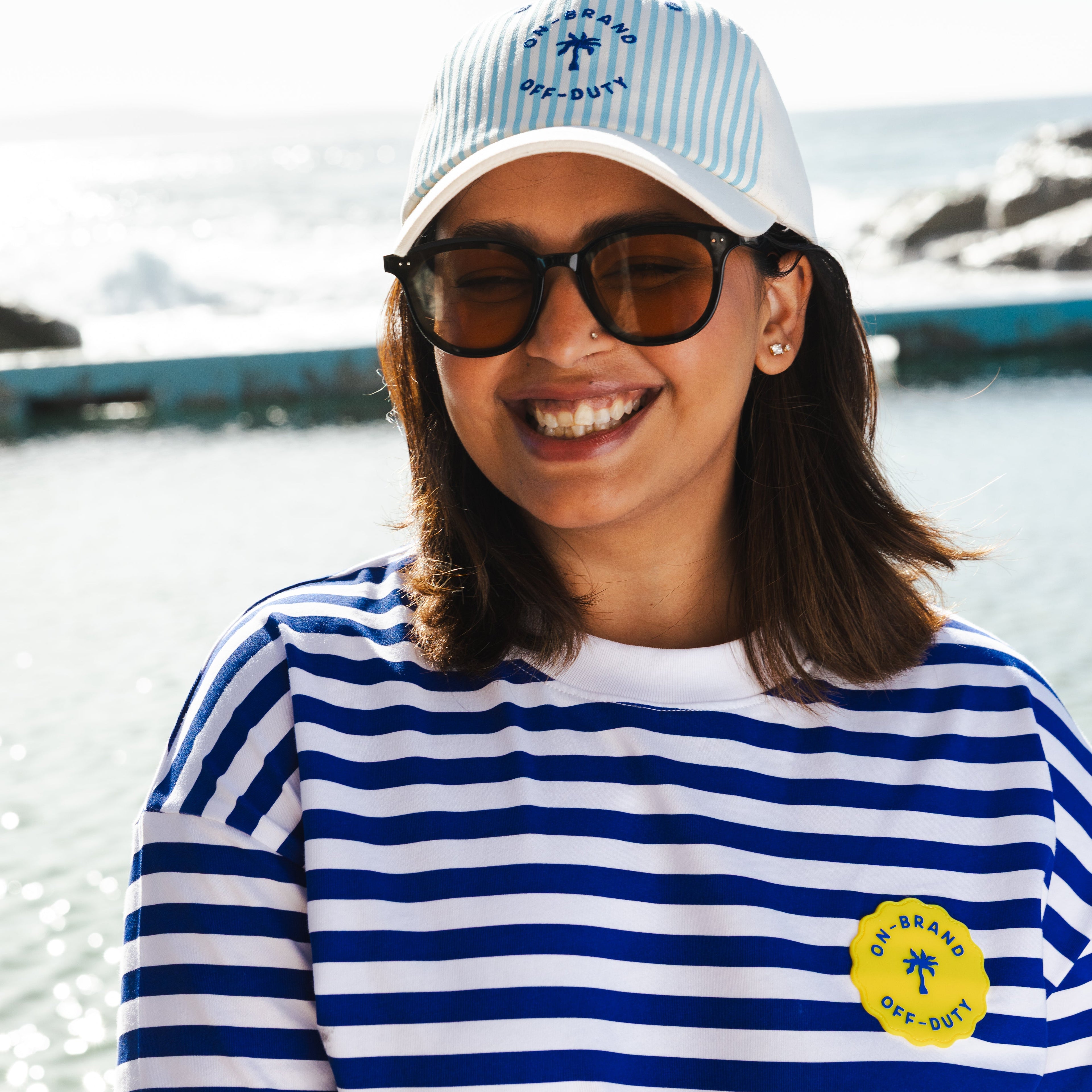 Person wearing a blue and white striped shirt with a cap and sunglasses, standing by water.