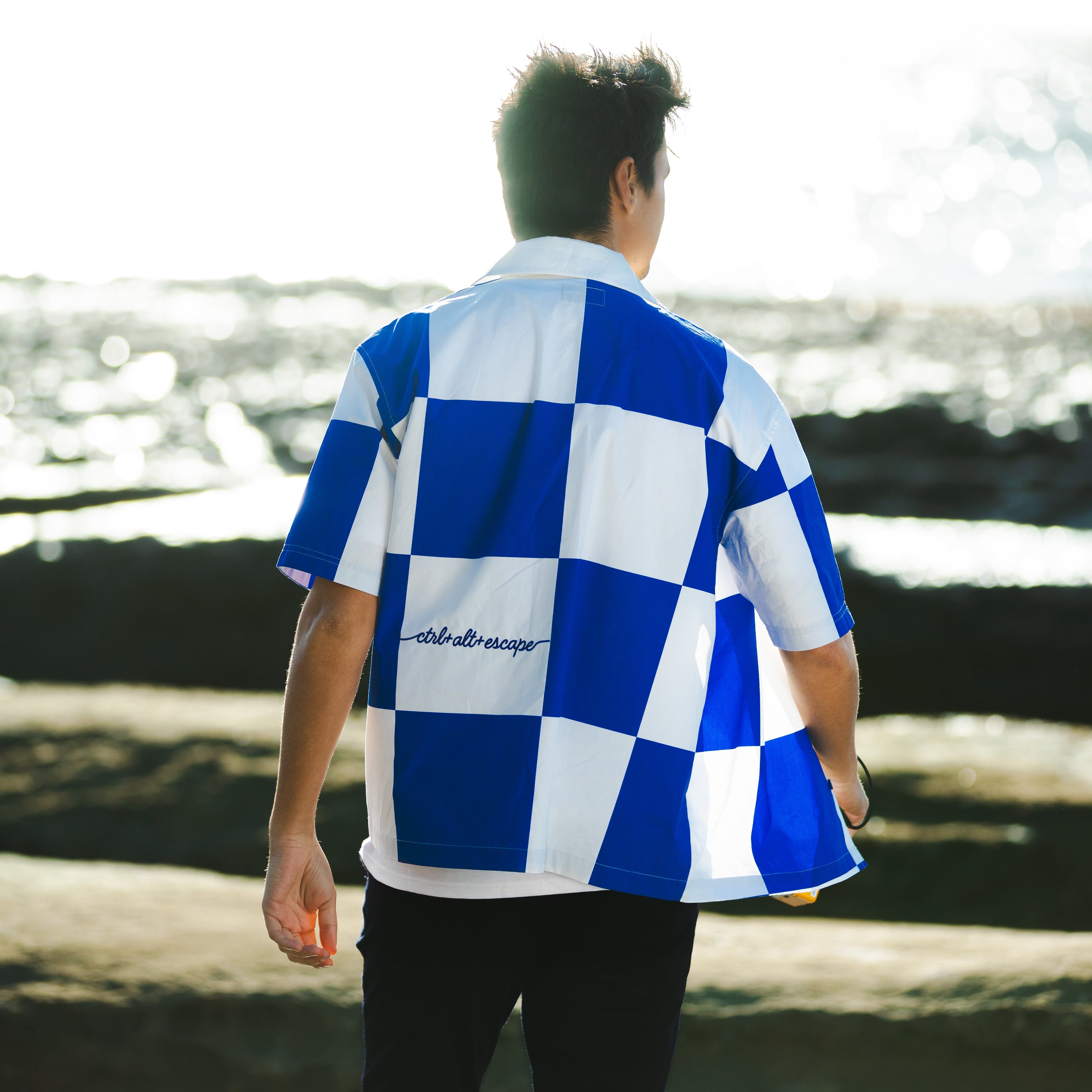 Person wearing a blue and white checkered shirt with text, standing on a beach.