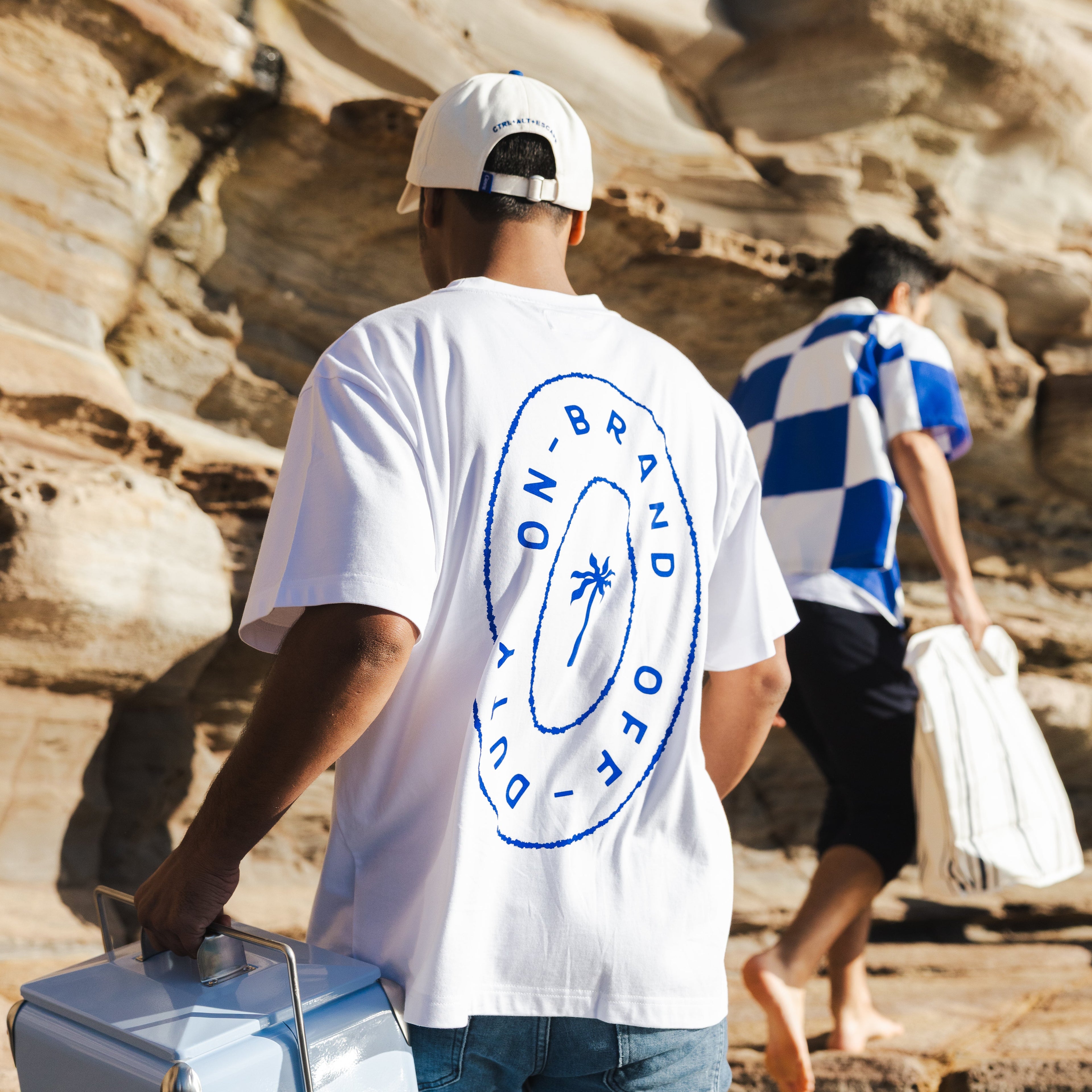 Two people walking on a rocky path with one carrying a cooler, wearing branded t-shirts.