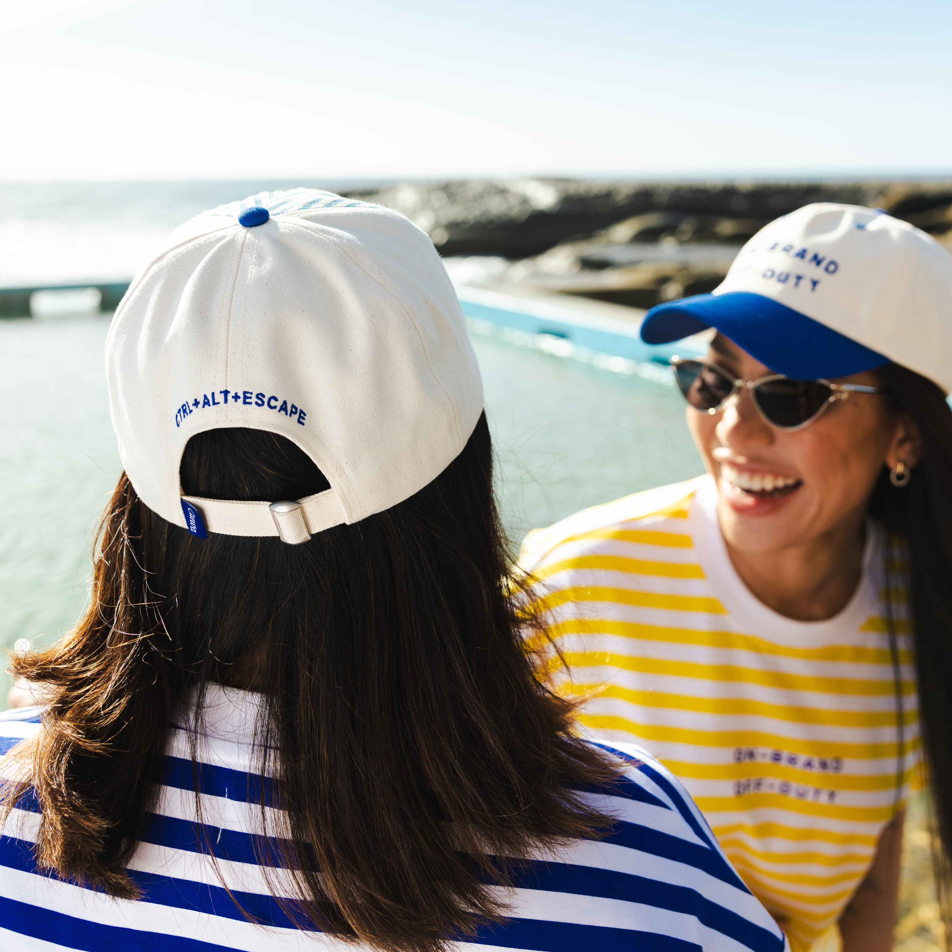 Two people wearing white caps with blue accents, one in a striped shirt and the other in a yellow and white striped shirt, standing on a beach.